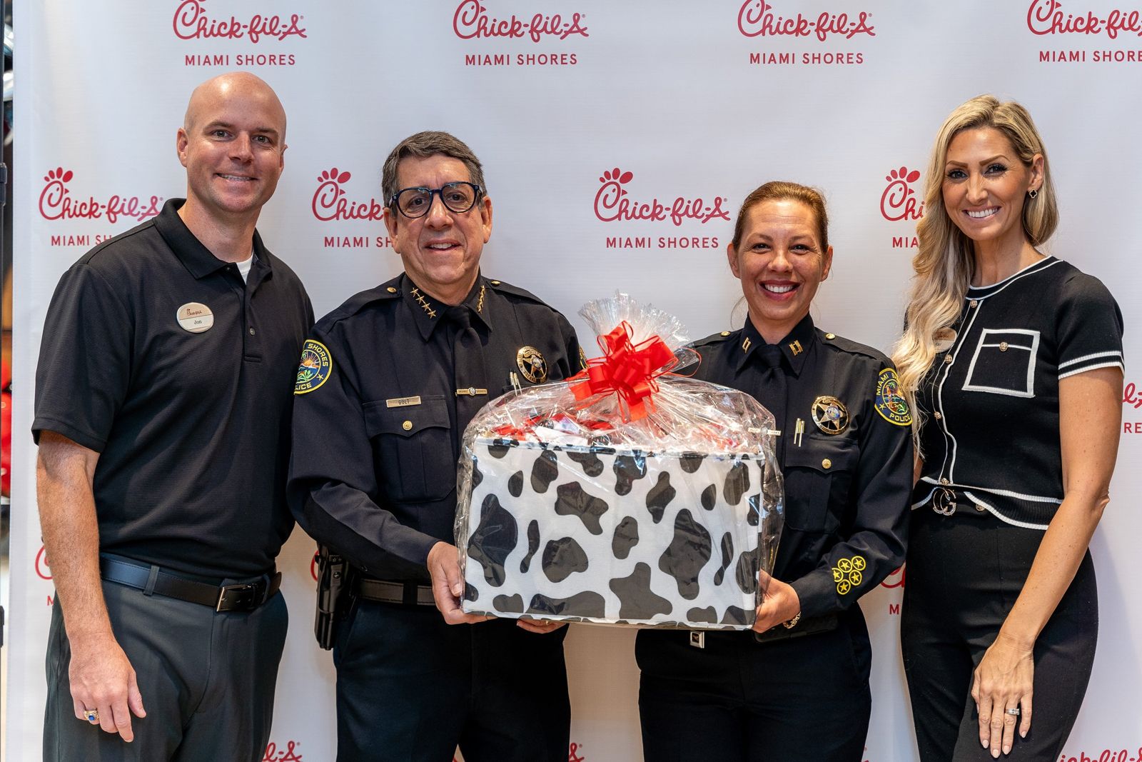 Two uniformed local police officers holding a cow-print gift basket with Chick-fil-A Miami Shores hosts at the grand opening