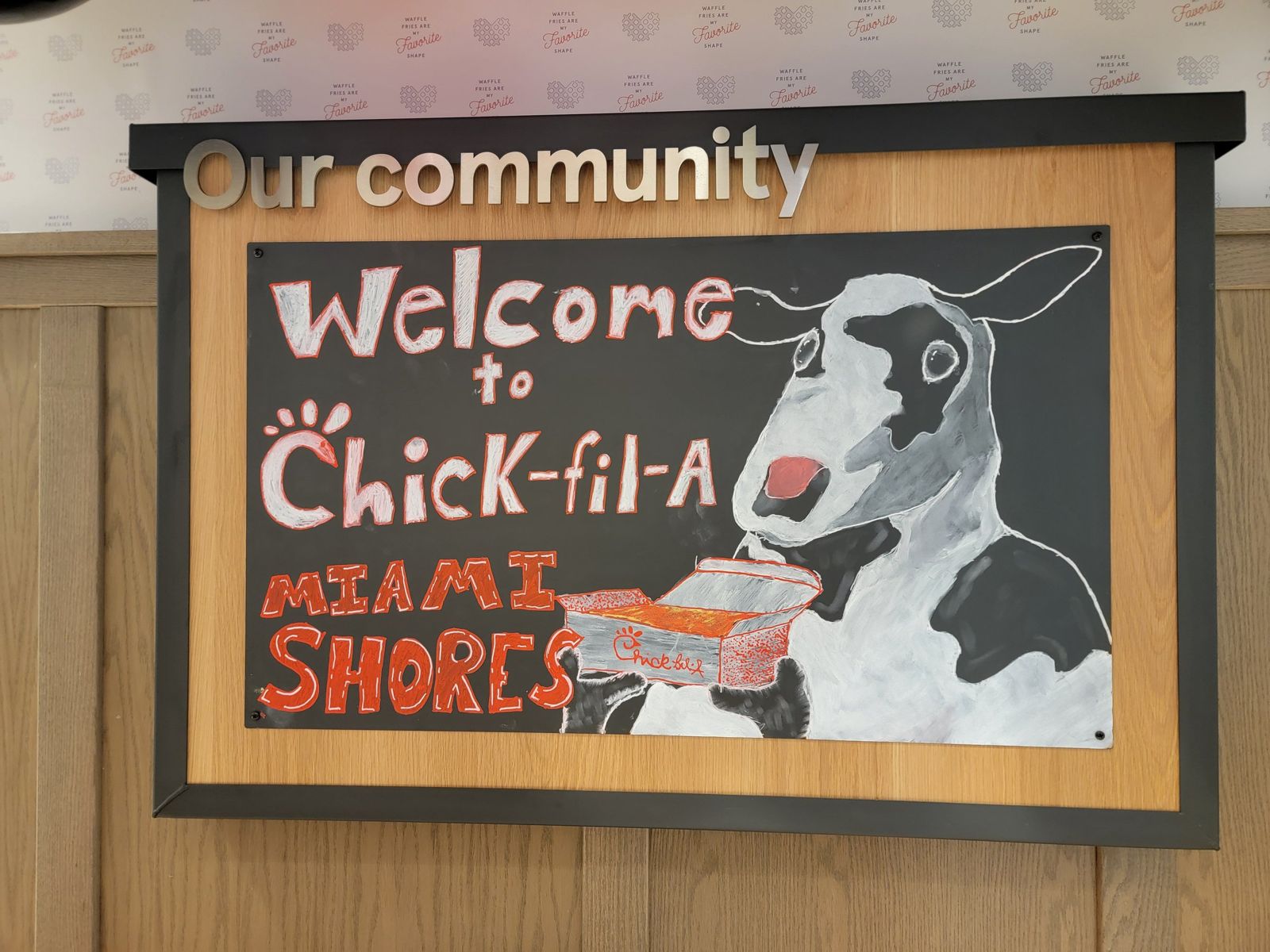 Hand-drawn chalkboard reading Welcome to Chick-fil-A Miami Shores, with a cow holding a nugget tray, under an Our community sign